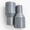 Dua fitting socket PVC Pralon berwarna abu berdampingan, satu bertuliskan Pralon dan satu bertuliskan 1 1/2" X 3/4", Socket 1 1/2 x 3/4 Inch TS Merek Pralon